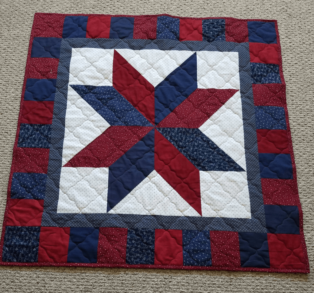 A handcrafted quilt featuring a star pattern in red, white, and blue fabrics, framed by a patchwork border.