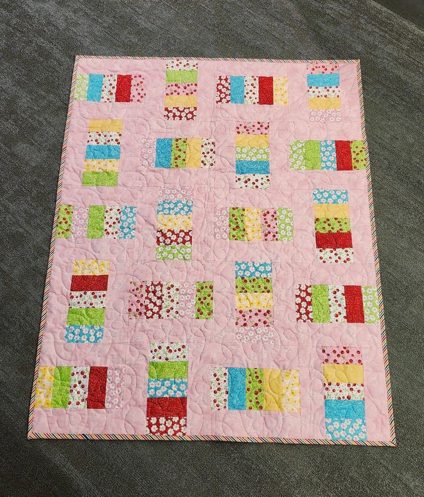 A colorful quilt featuring a patchwork design with various patterned blocks in red, green, blue, yellow, and pink on a light pink background.