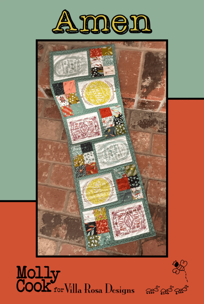 A colorful patchwork table runner featuring various fabric squares with text and designs, titled 'Amen' by Molly Cook for Villa Rosa Designs, set against a brick background.