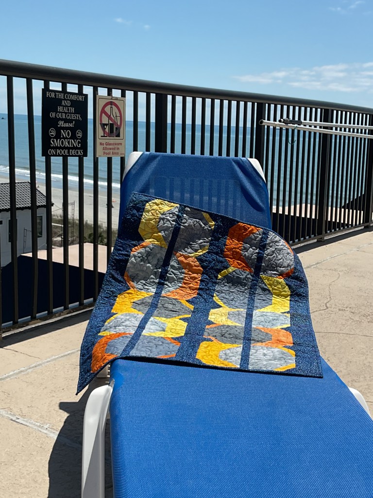 A colorful quilt draped over a blue lounge chair by a pool with ocean views and a no smoking sign in the background.