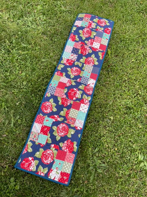 A colorful patchwork table runner featuring floral and geometric patterns, laid out on a grassy surface.