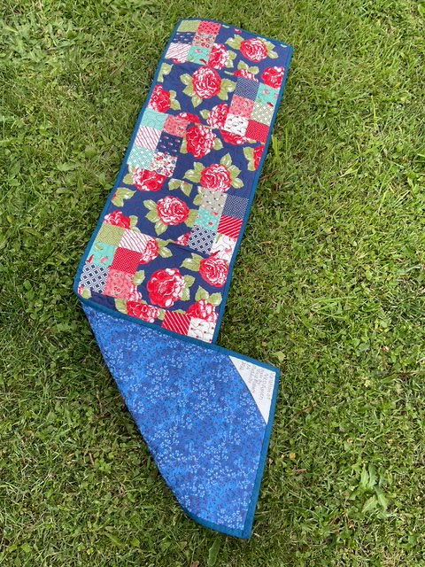 A colorful patchwork quilt featuring floral and geometric patterns, laid out on a grassy surface.
