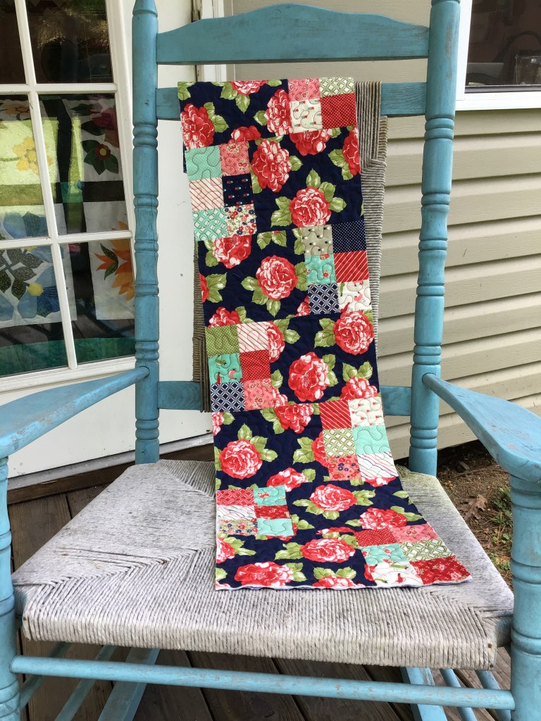 A colorful patchwork table runner featuring floral and geometric patterns, displayed on a blue wooden rocking chair.
