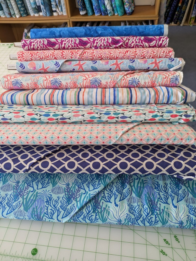 A collection of colorful fabric bolts stacked horizontally, featuring various ocean-themed patterns including coral, fish, and starfish. The background shows more fabric bolts arranged neatly.