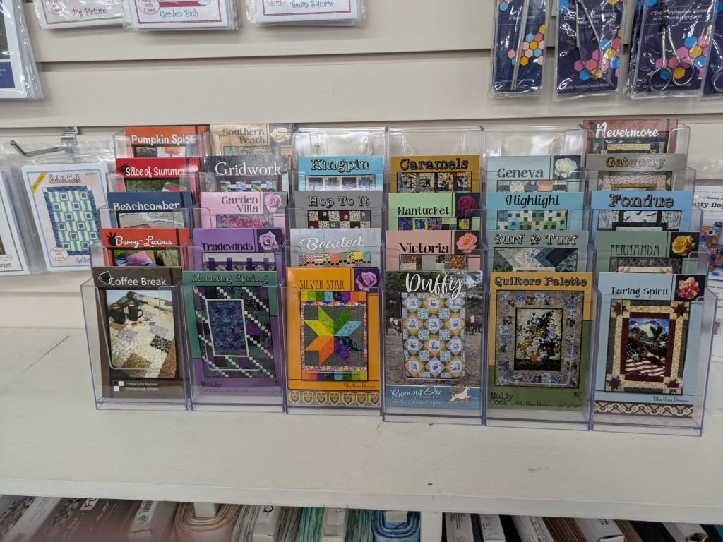 Display of various quilt patterns and designs shown in a retail setting, featuring colorful covers with titles such as 'Coffee Break,' 'Tradewinds,' and 'Silver Star' arranged in clear holders.