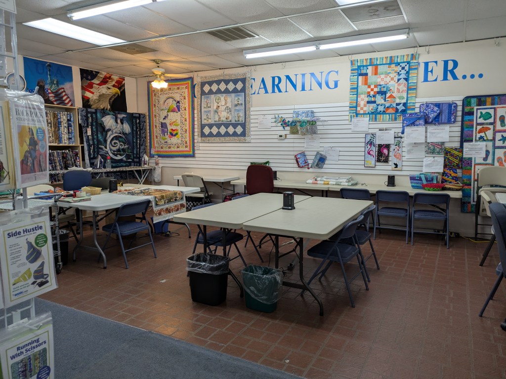 Interior of a craft studio with tables and chairs, featuring colorful wall hangings and quilt displays.