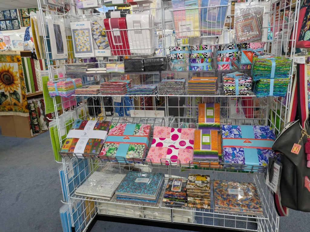 A display rack filled with various colorful fabrics and quilting supplies, showcasing organized stacks of patterned fabric, rolls, and packaged quilt kits in a craft store.