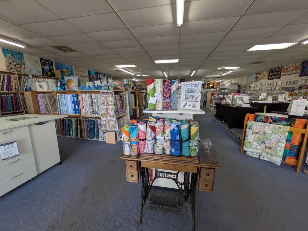 Interior of a fabric store featuring colorful fabric rolls on shelves, a central table with fabric bundles, and a variety of sewing supplies in the background.