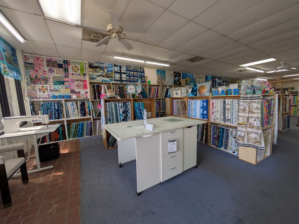 Interior view of a fabric store featuring numerous colorful fabric bolts, a cutting table, and sewing machines. Shelves display various quilting patterns and designs.