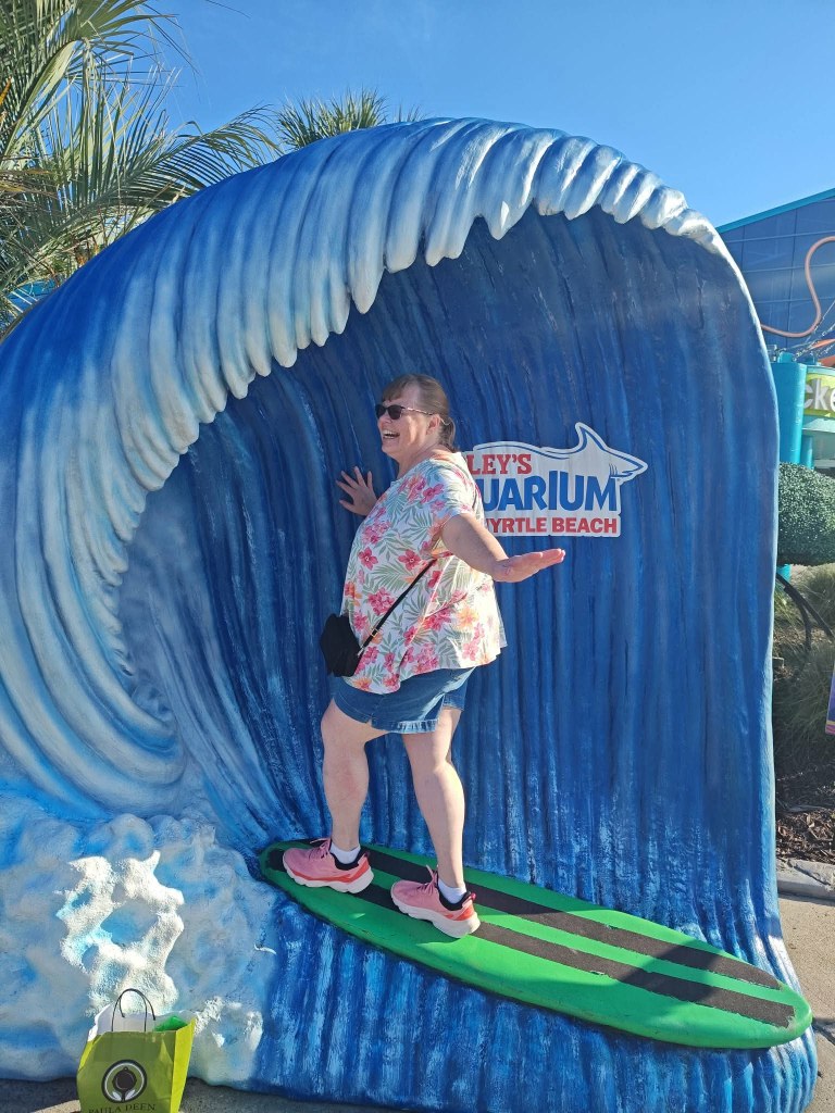 A woman standing on a surfboard in front of a large wave sculpture at Ripley's Aquarium in Myrtle Beach, smiling and posing with her arms outstretched.