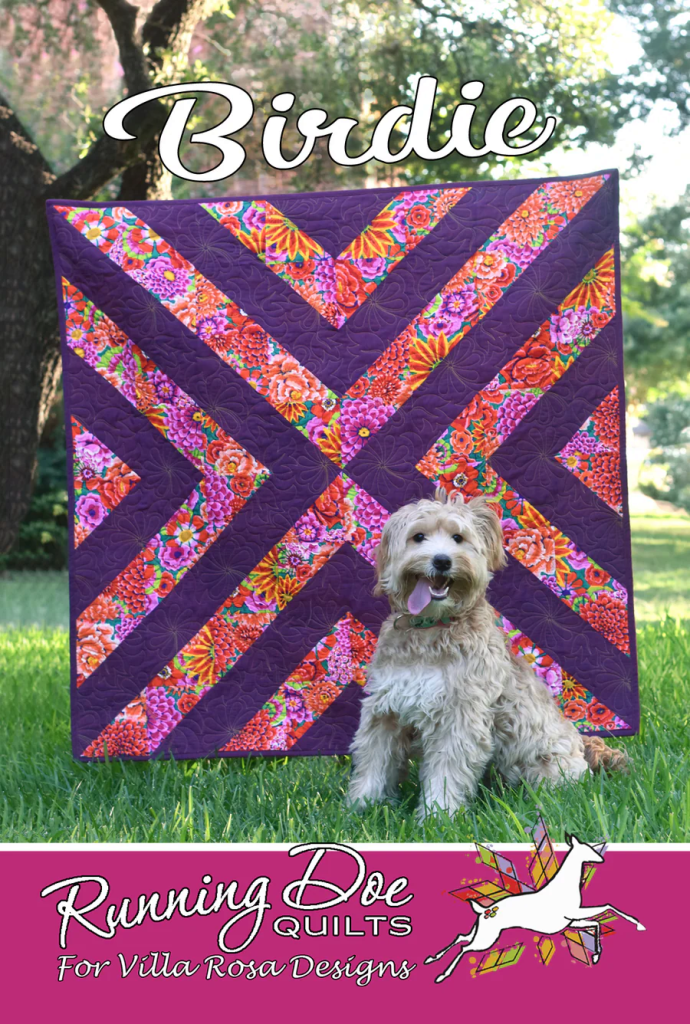 A fluffy dog sits in front of a vibrant quilt with purple and floral patterns, featuring the text 'Birdie' at the top and 'Running Doe Quilts For Villa Rosa Designs' at the bottom.