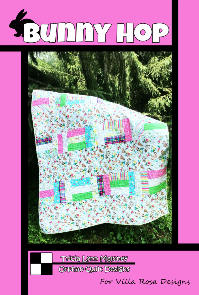 A colorful quilt titled 'Bunny Hop' displayed outdoors against a backdrop of green trees, featuring a mix of floral and geometric patterns.