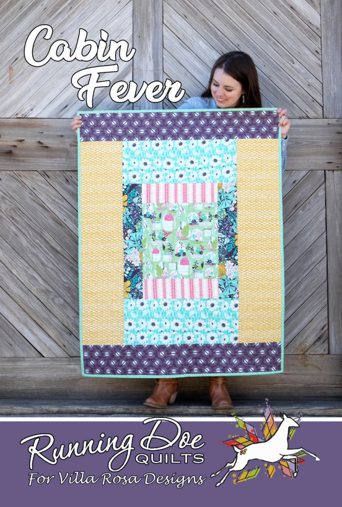 A woman holding a colorful quilt titled 'Cabin Fever' in front of a rustic wooden backdrop.
