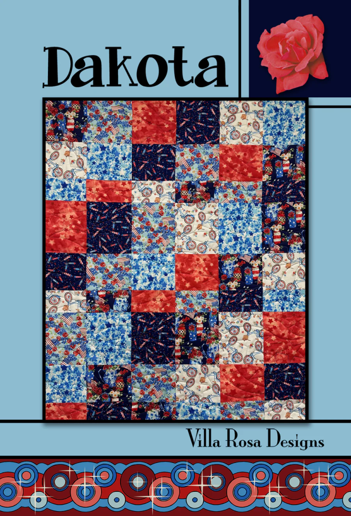 A colorful quilt featuring a patchwork design with red, blue, and cream fabrics, showcasing floral and festive patterns. The top of the image displays the word 'Dakota' and the brand 'Villa Rosa Designs'. A pink rose is also included in the design.