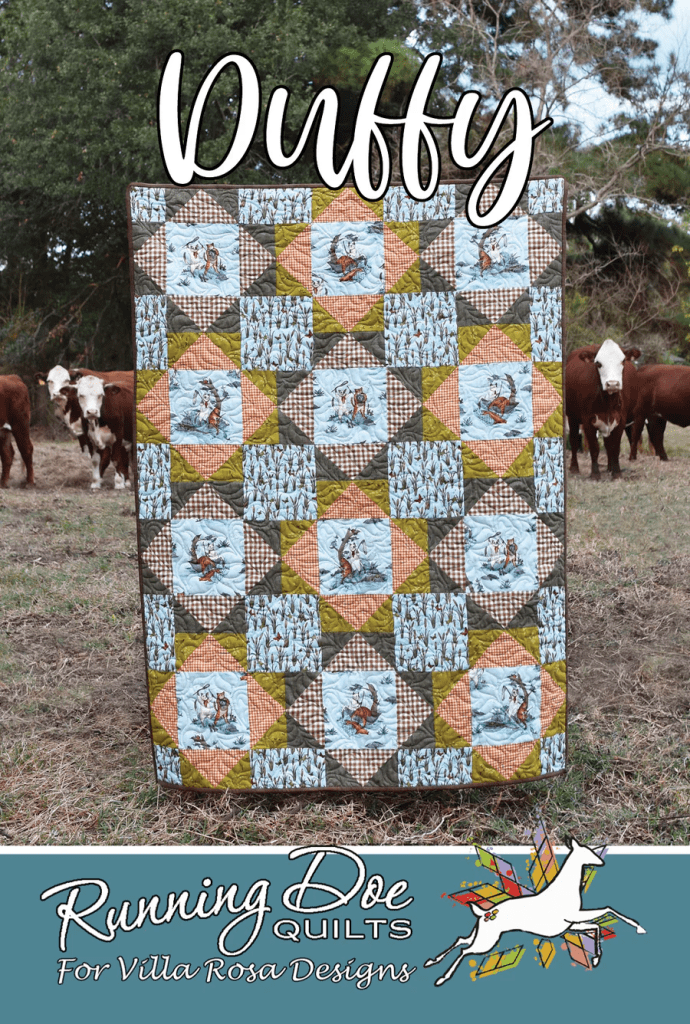 A colorful quilt named 'Duffy' displayed outdoors with cows in the background, featuring a mix of patterned squares and designs, under a blue sky.