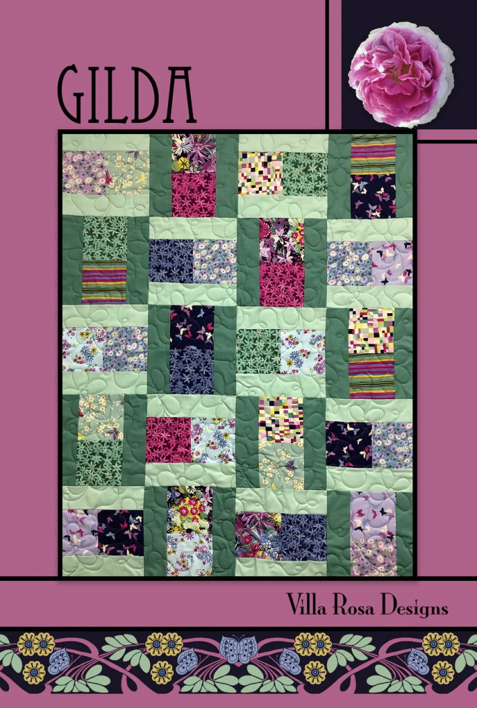 A colorful quilt design titled 'Gilda' by Villa Rosa Designs, featuring a patchwork pattern with various floral and geometric fabrics in shades of green, pink, and purple, set against a pink background.
