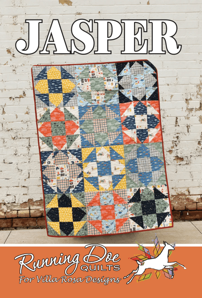 A colorful quilt named 'Jasper' displayed against a white brick wall, featuring geometric patterns in various shades of blue, red, yellow, and green. The quilt is partially draped over a flat surface, with a logo for Running Doe Quilts and Villa Rosa Designs at the bottom.
