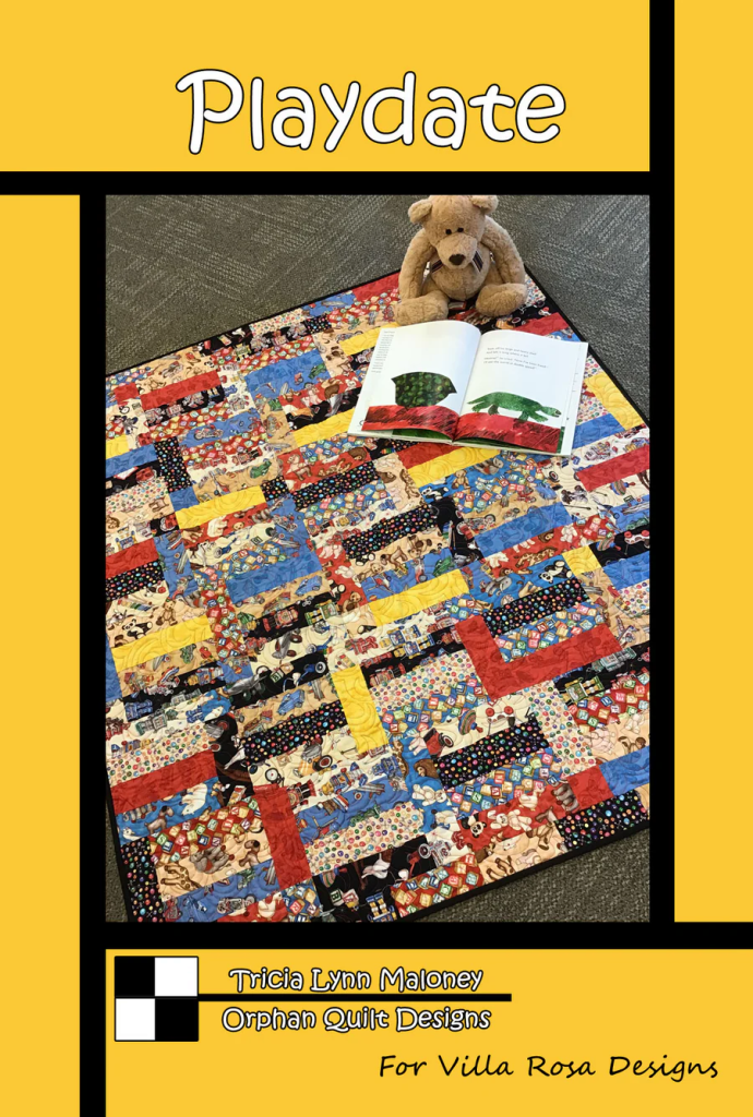 A colorful quilt titled 'Playdate' with various playful fabric patterns, placed on the floor with a stuffed bear and an open book on top.