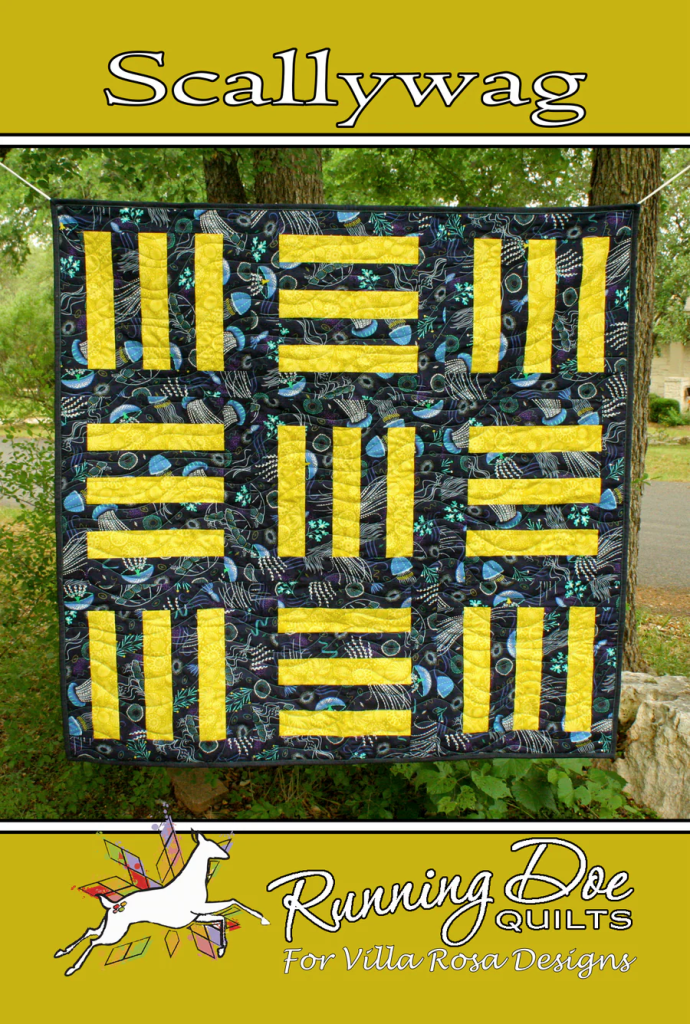 A colorful quilt named 'Scallywag' featuring a pattern of yellow and blue rectangular sections against a black background with abstract designs, displayed outdoors.
