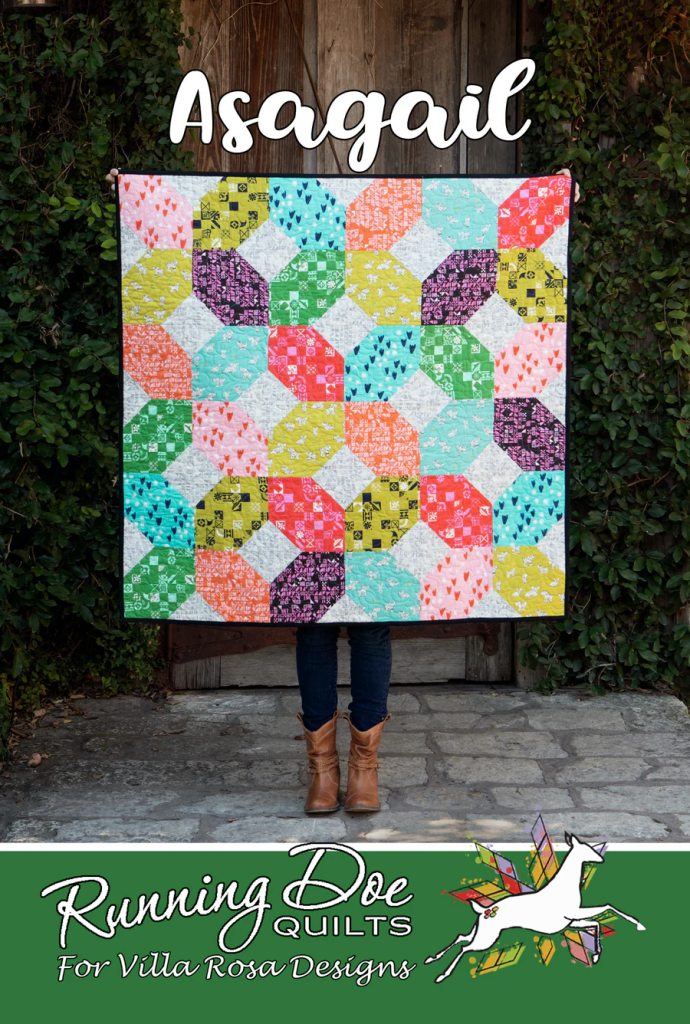 A person holding a colorful quilt named 'Asagail' featuring hexagonal patterns in various bright colors, standing against a wooden background with greenery.