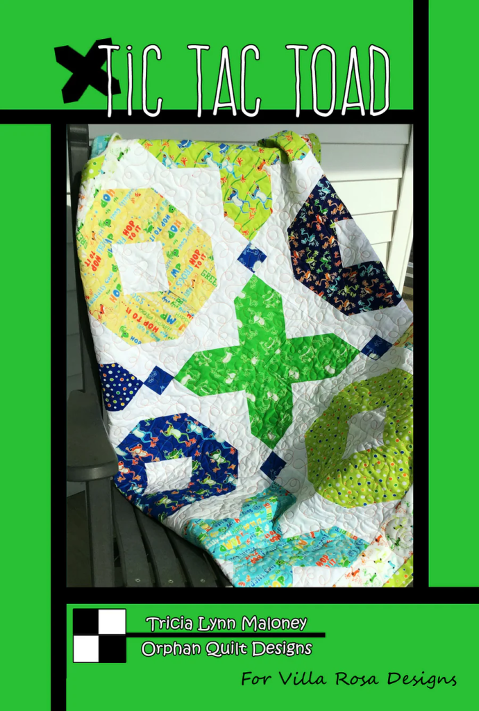 A colorful quilt titled 'Tic Tac Toad' featuring playful patterns and designs, displayed on a chair. The quilt is predominantly made of green, blue, yellow, and patterned fabrics.