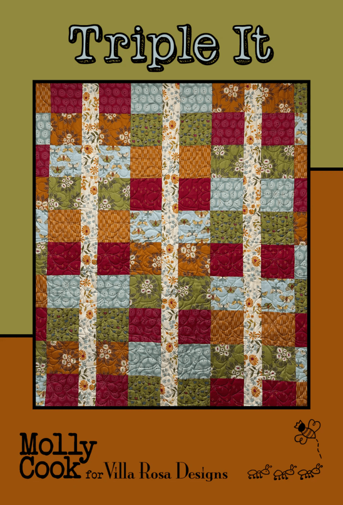 A quilt titled 'Triple It' designed by Molly Cook for Villa Rosa Designs, featuring a patchwork design with colorful fabrics in red, green, orange, and blue arranged in a grid pattern.