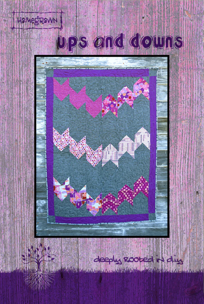 A colorful quilt titled 'Ups and Downs', featuring zigzag patterns in purple and pink tones set against a gray background. The quilt is framed with a purple border and displayed on a textured wooden background with pink hues.