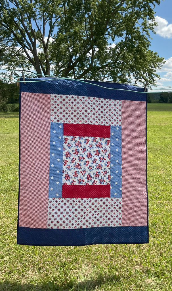 A colorful patchwork quilt featuring red, blue, and white fabrics with floral and star patterns, displayed outdoors against a grassy field with trees in the background.