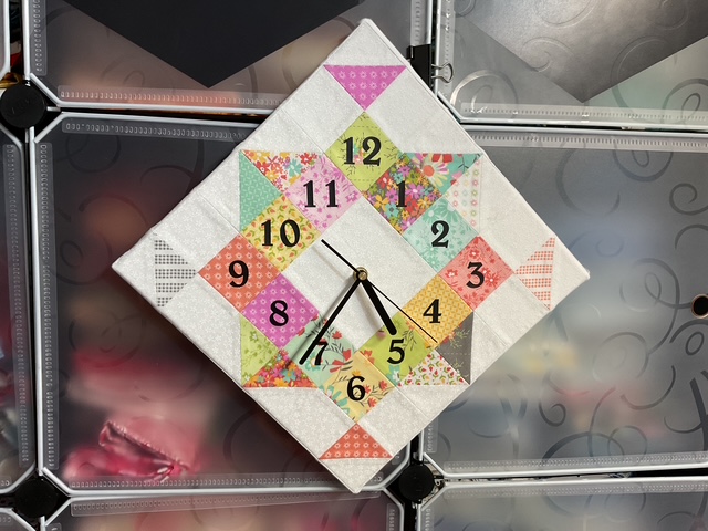A decorative wall clock with a diamond shape, featuring colorful patchwork patterns and black hour and minute hands, displayed against a patterned background.