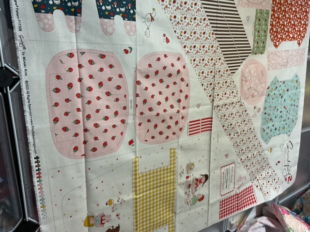 A fabric print featuring various colorful patterns including strawberries, polka dots, and floral designs, along with sewing patterns and illustrations of small characters.