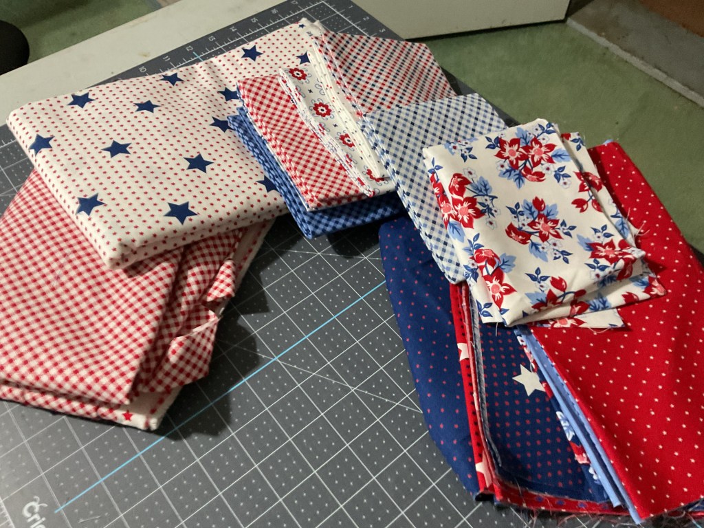 A collection of folded fabric pieces featuring various patterns, including polka dots and stars, in red, white, and blue colors, arranged on a cutting mat.