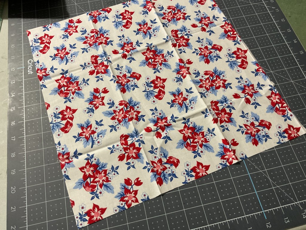 A square piece of fabric with a floral pattern featuring red, blue, and white flowers on a light background, placed on a cutting mat.