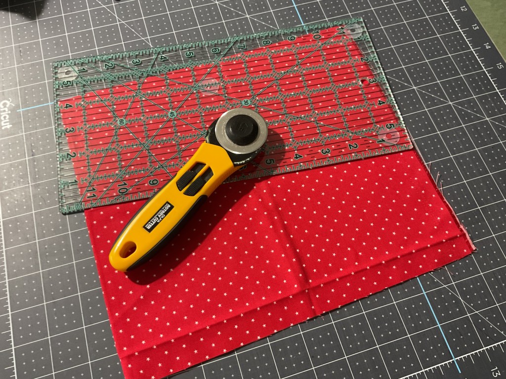 A rotary cutter positioned on a red polka dot fabric square, with a transparent ruler placed over it, resting on a cutting mat with grid lines.