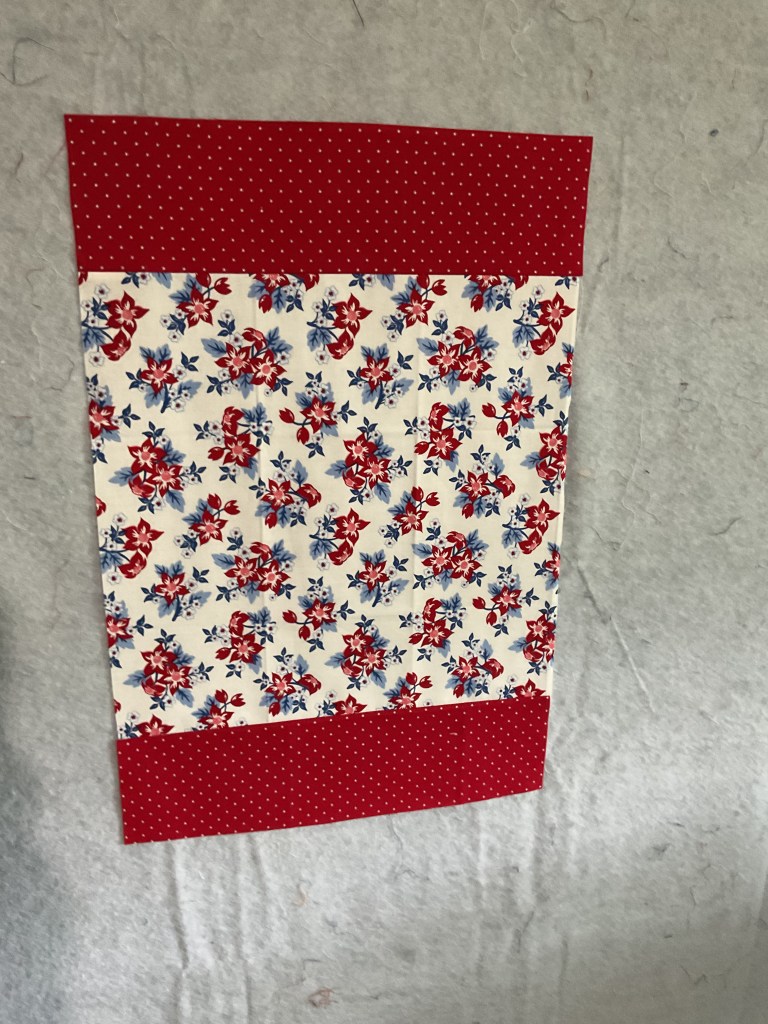 A patchwork fabric piece featuring a floral pattern with red and blue flowers on a cream background, bordered at the top and bottom with solid red fabric with white polka dots.