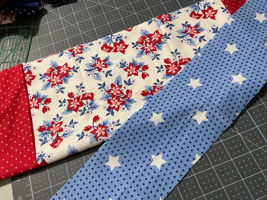 An arrangement of red, white, and blue fabric pieces, featuring floral patterns and star designs, placed on a cutting mat.