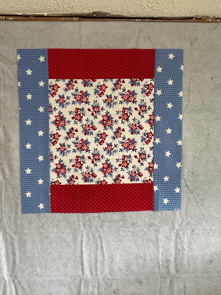 A quilt square featuring a floral print center surrounded by red and blue patterned borders with polka dots and stars.