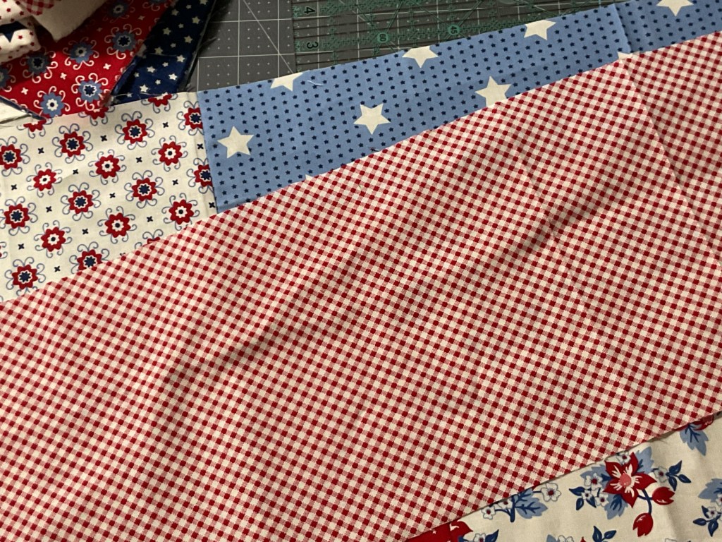 A close-up of colorful fabric squares, featuring a mix of red and blue patterns including checks, floral designs, and stars, laid out on a cutting mat.