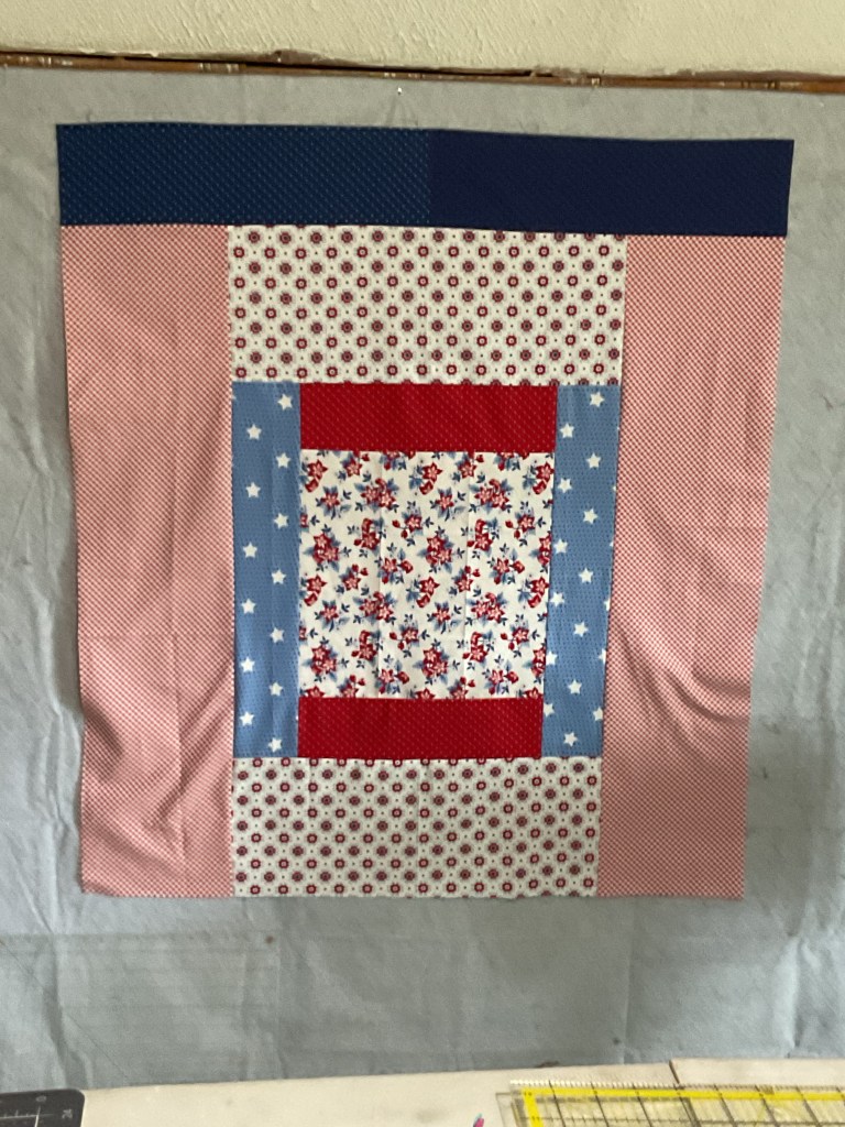 A colorful quilt top featuring a central floral pattern, surrounded by varying patterns of red, blue, and pink fabric.