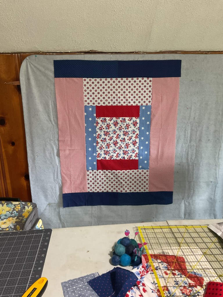 A handmade quilt displayed on a wall, featuring a patchwork design with red, white, and blue fabrics, surrounded by a blue border.