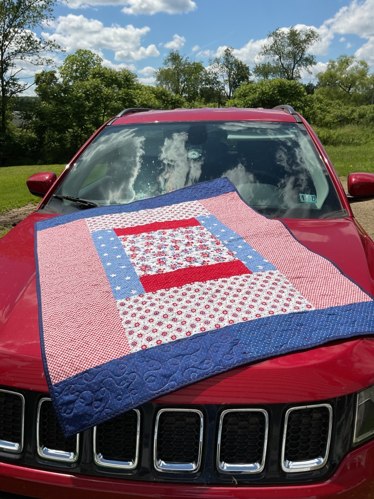 A colorful quilt displayed on the hood of a red car under a blue sky with fluffy clouds and green trees in the background.