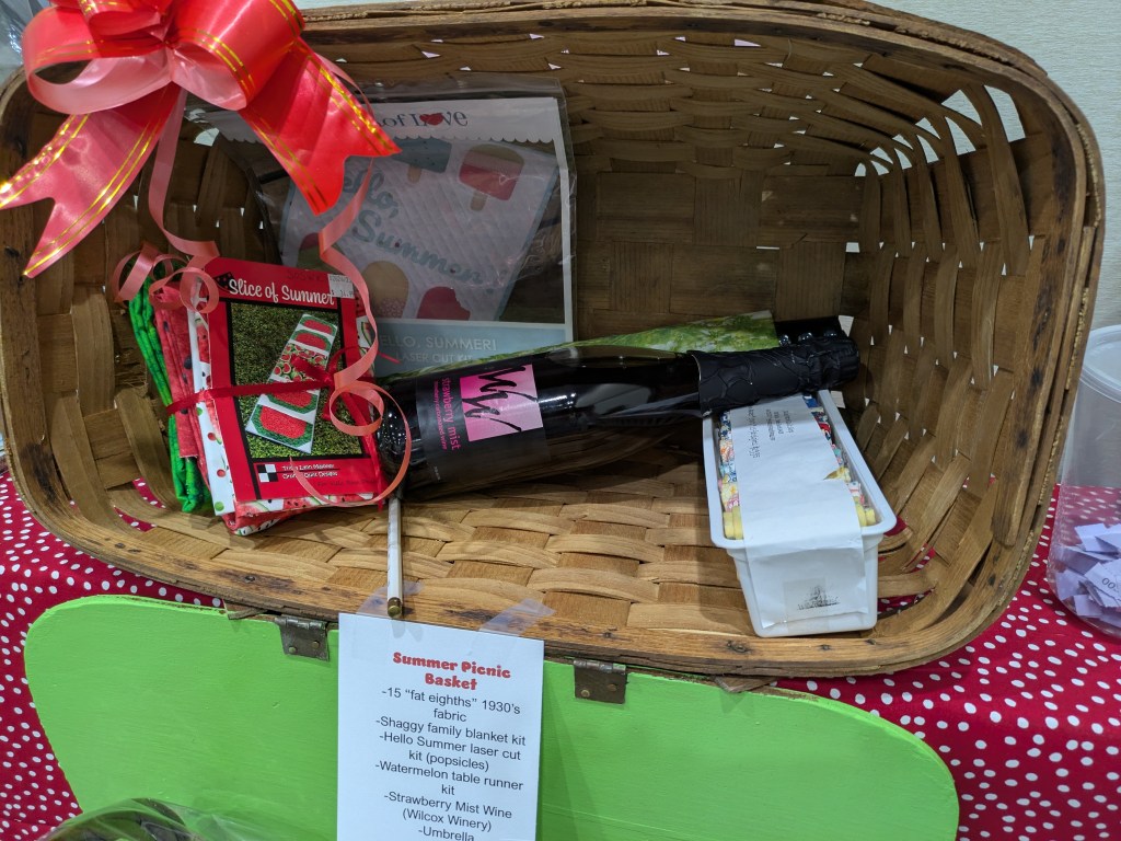 A wicker basket filled with summer picnic items, including fabric squares, a bottle of wine, and craft kits for a table runner and popsicles, topped with a red ribbon.