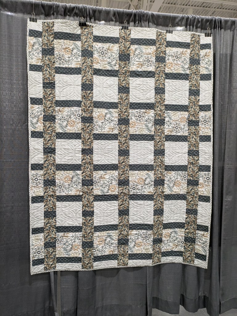 A quilt displayed on a hangar, featuring a pattern of alternating squares with floral designs and dark stripes, showcasing intricate stitching.