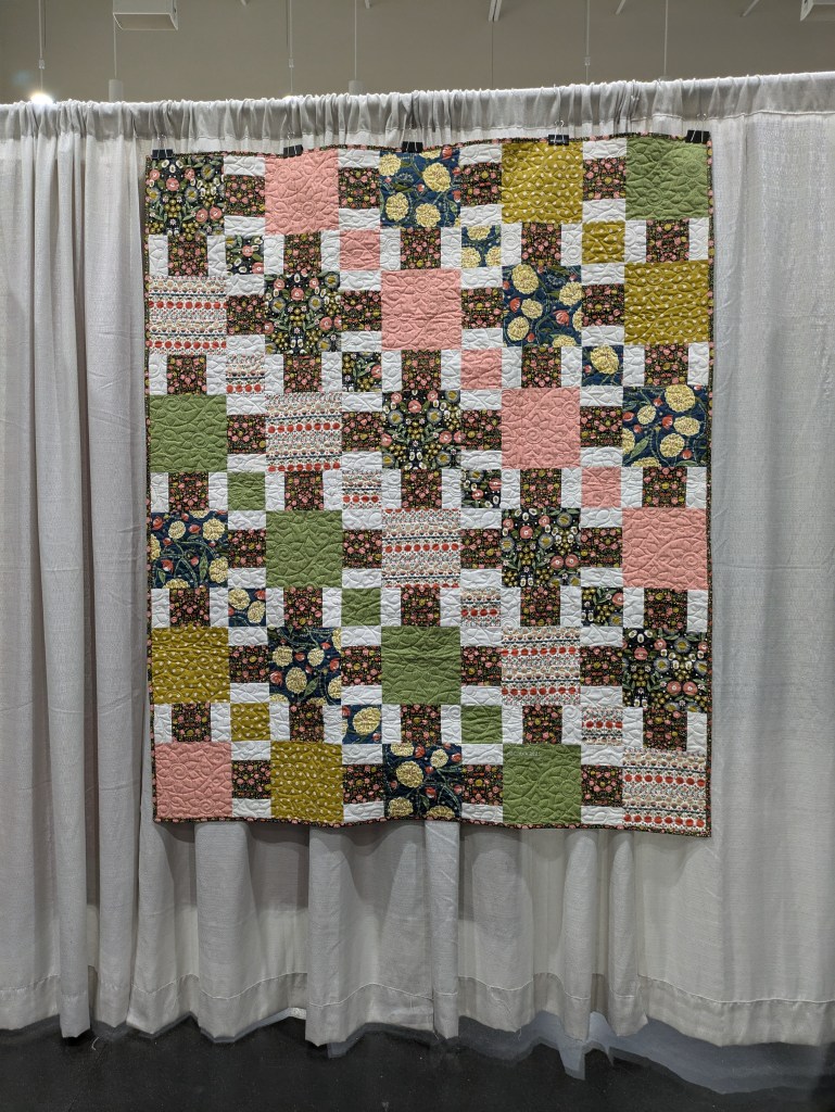 A colorful quilt displayed on a wall, featuring a patchwork design with floral and geometric patterns in shades of pink, green, yellow, and white.