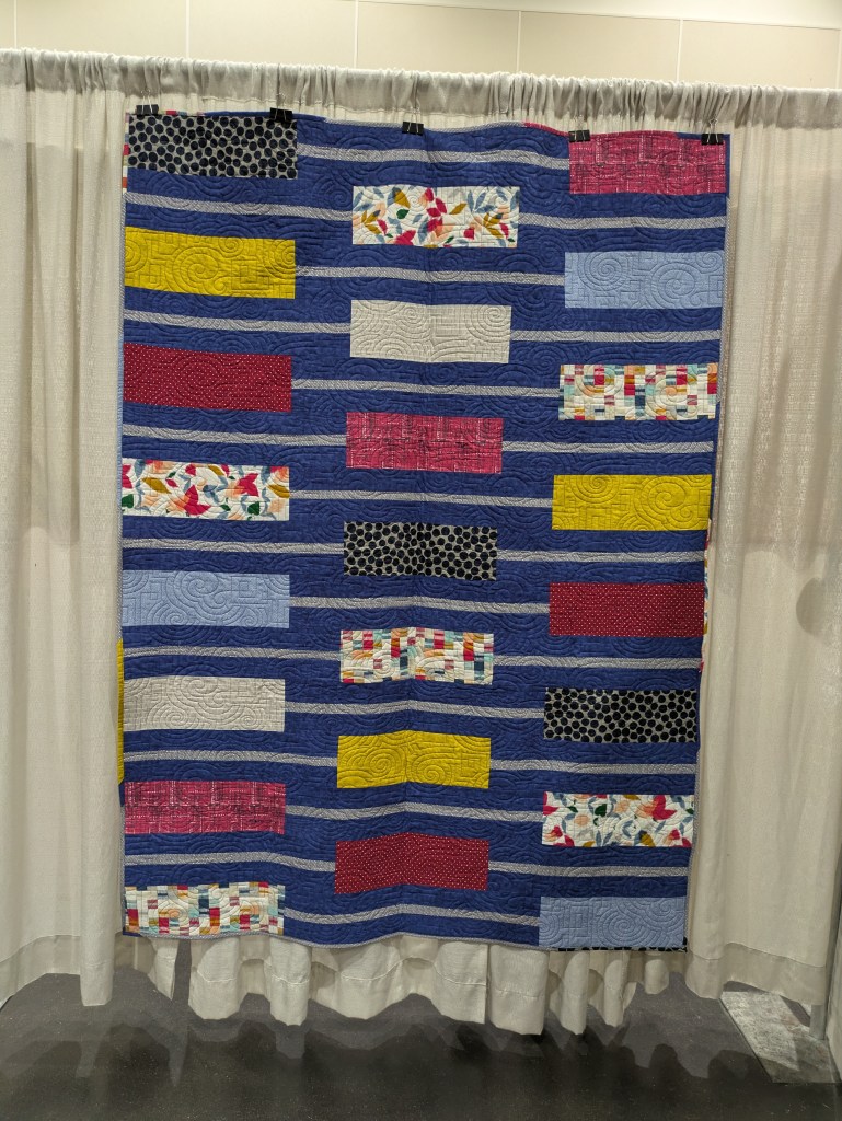 A colorful quilt featuring horizontal stripes in various patterns and colors, displayed against a backdrop of light fabric.