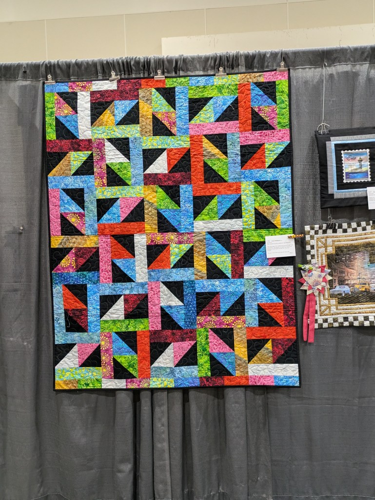 Colorful quilt displayed on a gray backdrop, featuring a geometric pattern of triangles in various vibrant colors including red, blue, green, pink, and black.