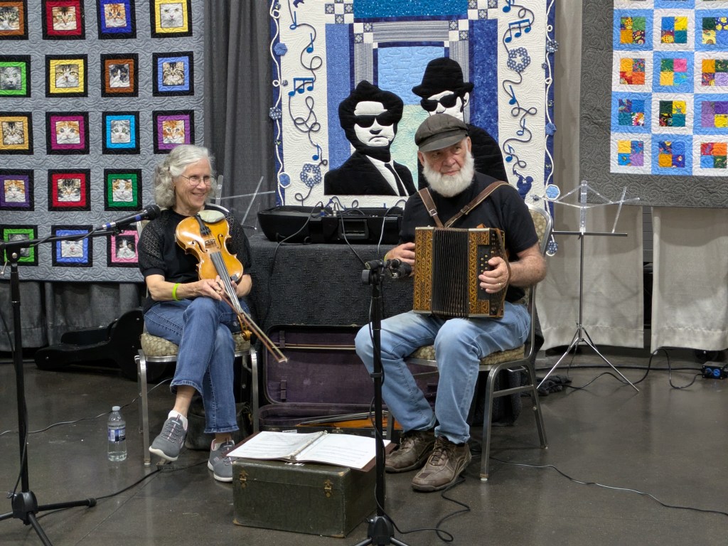 A woman playing the violin and a man playing an accordion on stage, with vibrant quilts featuring various designs in the background.