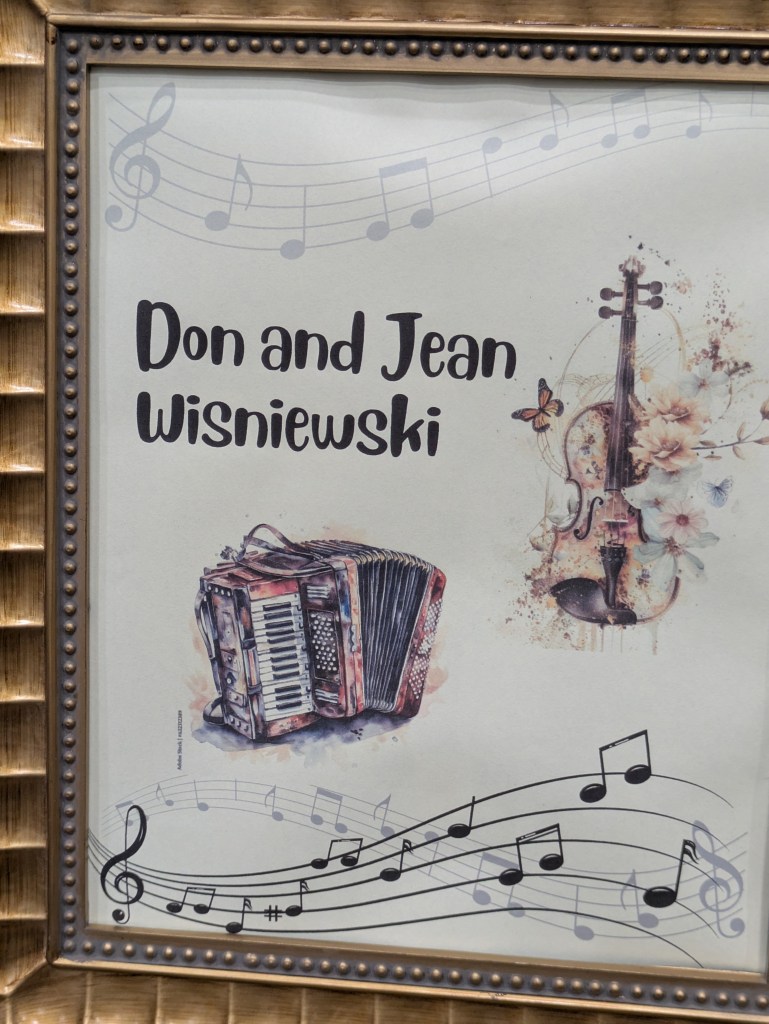 A decorated frame featuring the names 'Don and Jean Wisniewski' accompanied by illustrations of a violin and an accordion, with musical notes in the background.