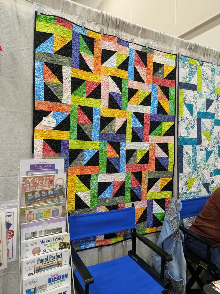 Colorful quilt display featuring a geometric pattern with bright colors on a black background, alongside a blue and white quilt in a craft booth setting.