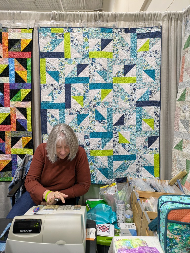 A woman with long gray hair seated at a table with a cash register, working in a booth surrounded by colorful quilts displayed behind her.