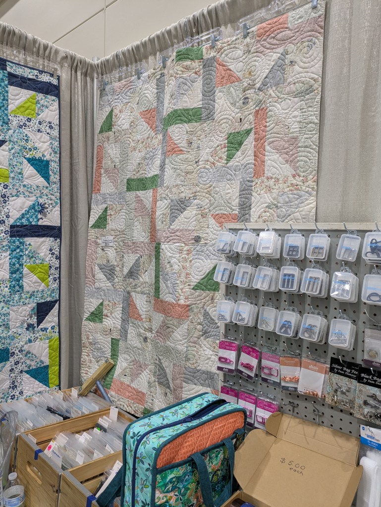 A display at a fabric show featuring colorful quilts hanging on the wall and various quilting supplies organized on a pegboard. The quilts include geometric patterns in pastel and bold colors, while the supplies include quilting tools and accessories.